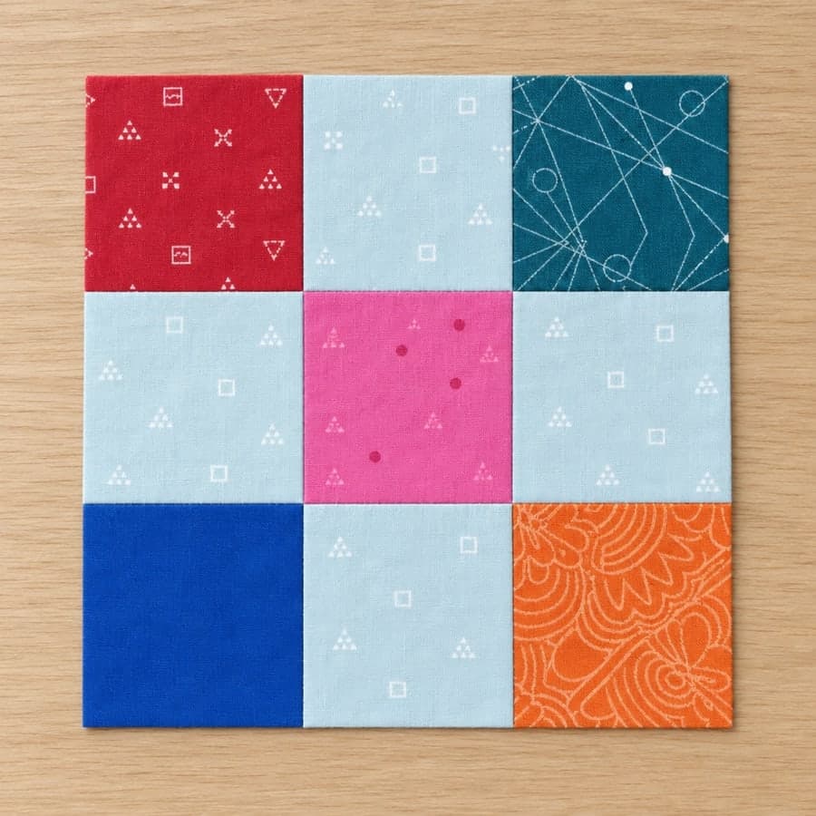 One finished nine-patch quilt block on a pale oak board: four red corners and a hot pink centre on a pale ice-blue print.