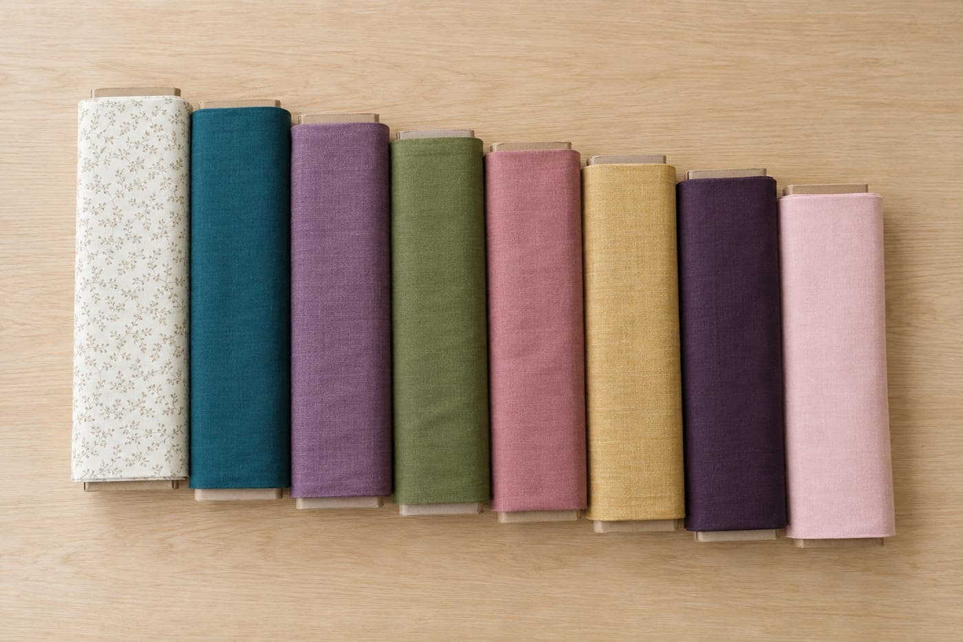Eight bolts of quilting cotton lying in a row on a pale oak board — cream, teal, plum, olive, dusty rose, gold, aubergine and pink.