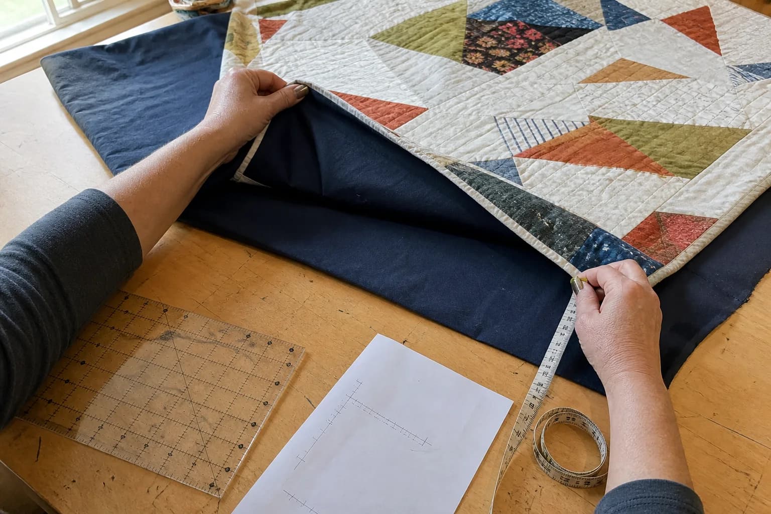 A quilter lifts a modern patchwork quilt top to measure the extra navy backing fabric around its edges on a used wooden table.