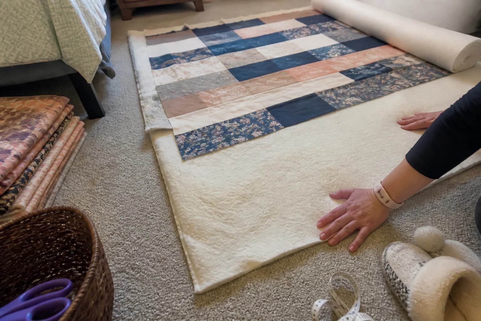 A quilter measures batting from a partly opened roll beside a quilt top in an ordinary sewing room.