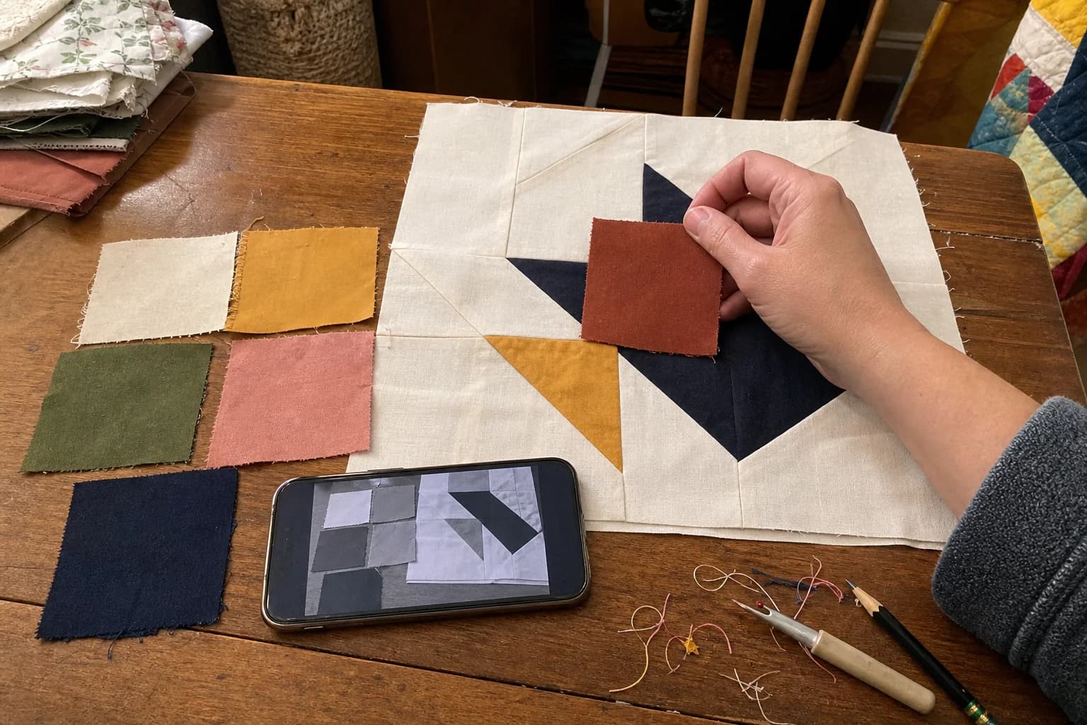 A quilter auditions a rust fabric swatch beside cream and navy fabrics while a phone shows the same quilt block in grayscale.