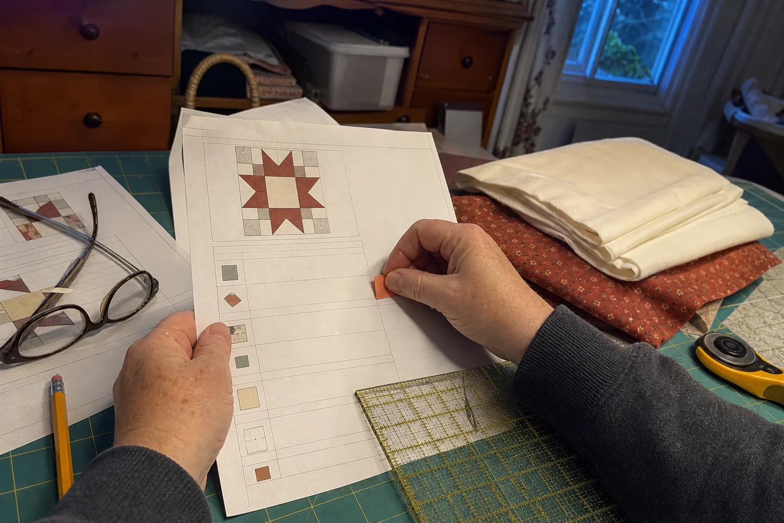 A quilter checks printed pattern pages beside folded cream and rust fabrics, a ruler, pencil, and reading glasses at a home cutting mat.
