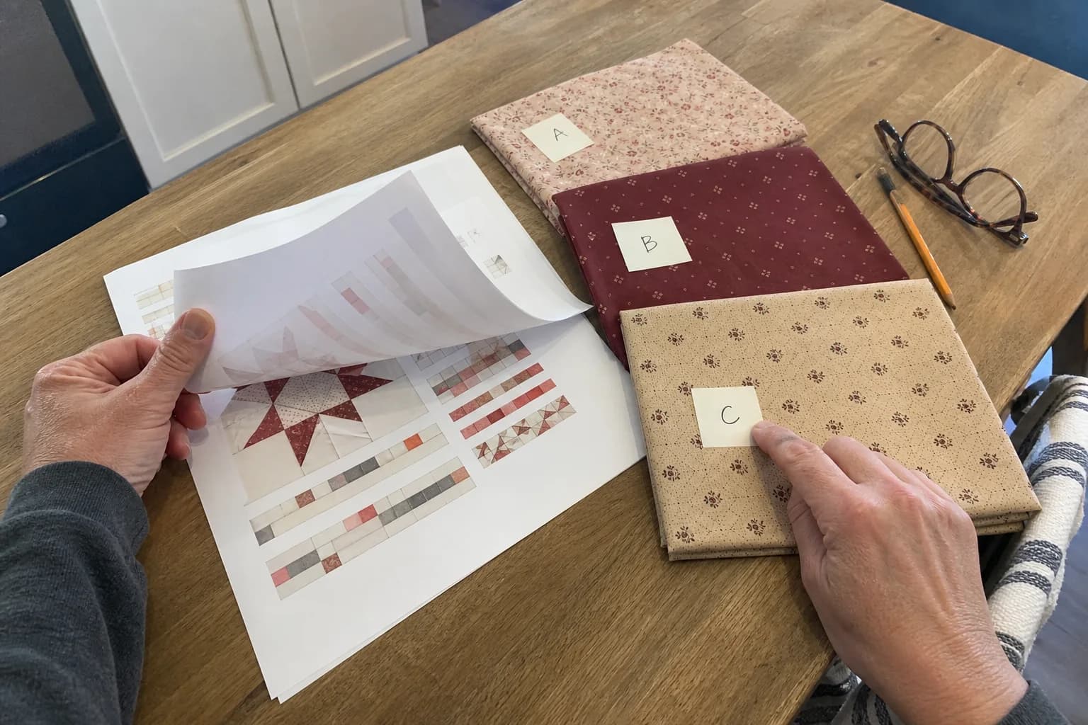 A printed quilt pattern, labeled fabric swatches, pencil notes, and a ruler on a busy cutting table.