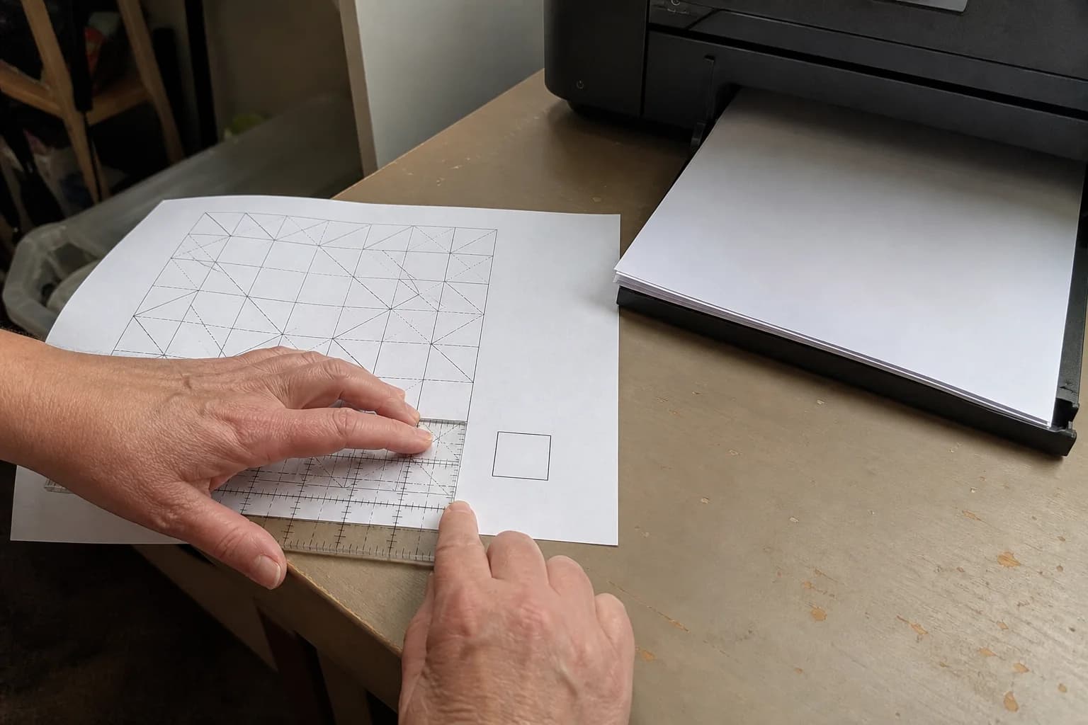 A quilter measures a printed calibration square with an acrylic ruler beside a basic home printer.